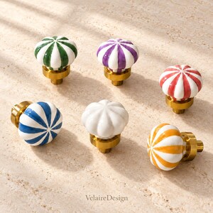 May include: Six ceramic cabinet knobs with a fluted, pumpkin-like shape and a brass-toned metal base. The knobs feature vertical stripes in green, purple, red, blue, white, and yellow. The text VelaireDesign is printed at the bottom.