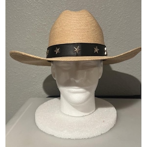 Könnte beinhalten: Ein hellbrauner Cowboyhut aus Stroh mit einem schwarzen Band um die Krone. Das Band ist mit gleichmäßig verteilten, silberfarbenen Metallsternen verziert. Der Hut hat eine strukturierte Krempe, die an den Seiten nach oben gebogen ist.