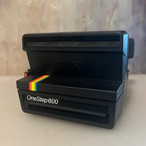 May include: A black Polaroid OneStep 600 instant camera featuring a prominent rectangular body, a fixed lens housing, and a signature rainbow-colored stripe accent on the front panel. The text OneStep 600 is printed in white on the lower front.