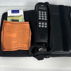 May include: A black GTE cellular telephone with a numeric keypad and function buttons, housed in a black carrying case. Included are a Motorola owner's manual, a programming instruction card, and an orange Motorola limited warranty document.