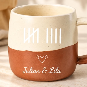 Può includere: Una tazza in ceramica bicolore con la parte superiore color crema e la base color terracotta. La tazza presenta un grafico con segni di conteggio bianchi, una piccola icona a forma di cuore e il testo "Julian & Lila" stampato in bianco.