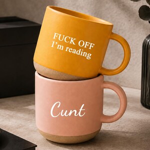 May include: Two stacked ceramic mugs with a matte finish. The top mug is mustard yellow with the white text "FUCK OFF I'm reading" and the bottom mug is dusty pink with the white text "Cunt" in a cursive font. Both mugs have a natural base.