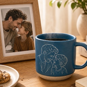 May include: A matte blue ceramic mug featuring a white line art illustration of a person and a child looking at each other. The mug has a textured, speckled light gray base and a curved handle, with steam rising from the dark liquid inside.
