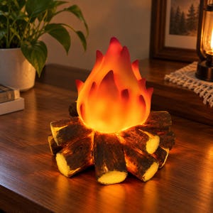 May include: A decorative lamp shaped like a campfire, featuring a translucent, flame-coloured top in shades of orange, yellow, and red, resting on a base of textured, brown, log-shaped pieces that emit a warm, glowing light from within.