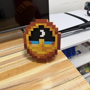 Op de afbeelding: Een cirkelvormig pixel art object met een bruine buitenrand, een zwart bovengedeelte met een wit maansikkel-symbool, en een ondergedeelte met een geel en oranje kleurverloop en twee horizontale blauwe rechthoekige accenten.