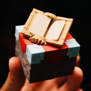May include: A small, cubic, 3D-printed object featuring a dark brown base with light blue corner accents and a red top surface. An open, tan-coloured book with two blank pages is positioned on top of the cube, creating a blocky, pixelated aesthetic.