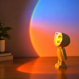 Könnte beinhalten: Eine weiße, roboterförmige Projektionslampe mit rundem Kopf und einer hellgelben Lichtquelle. Die Lampe wirft einen großen, lebendigen, sonnenuntergangsartigen Bogen aus orangefarbenem, rosa und blauem Licht an die Wand und erzeugt einen Farbverlauf.
