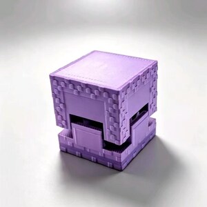Könnte beinhalten: Ein hellviolettes, würfelförmiges Objekt mit einer pixeligen Textur und einem deutlichen horizontalen Spalt, der den oberen Teil von der Basis trennt. Das Design weist ein blockartiges Gittermuster an den Kanten auf, was eine geometrische Form ergibt.