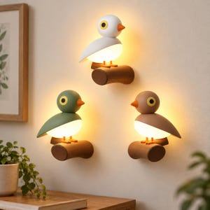 Puede incluir: Tres lámparas de pared con forma de pájaro, cada una posada sobre una rama cilíndrica marrón. Los pájaros son blancos, verde salvia y marrones, con picos naranjas, ojos negros con borde amarillo y vientres blancos que emiten luz cálida.