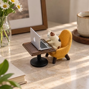 Puede incluir: Una figura en miniatura de un gato blanco y marrón sentado en un sillón amarillo texturizado frente a una pequeña mesa de madera marrón oscuro. El gato está colocado frente a un portátil plateado con un logo de apple visible en la tapa.