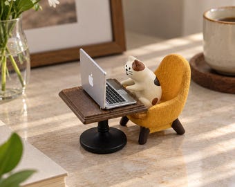 May include: A miniature figurine of a white and brown cat sitting in a textured yellow armchair at a small dark brown wooden table. The cat is positioned in front of a silver laptop with a visible apple logo on the lid and a black keyboard.