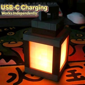 Könnte beinhalten: Eine quadratische, blockförmige Laterne mit einem dunkelgrauen Rahmen und einem leuchtend orangefarbenen Zentrum. Die Oberseite verfügt über einen Griff. Der Text über der Laterne lautet: USB-C Charging, Works Independently. Das Design imitiert eine Pixel-Optik.