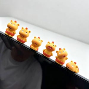 Könnte beinhalten: Eine Reihe von sechs kleinen gelben Nilpferd-Figuren mit orangefarbenen Shorts, jede mit einem einzigartigen Gesichtsausdruck, die auf dem oberen Rand eines schwarzen Monitorrahmens sitzen. Die Figuren haben abgerundete Ohren.