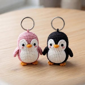 Può includere: Due portachiavi a forma di pinguino all'uncinetto, uno rosa e uno nero, ciascuno con pancia bianca, becco arancione, piedi arancioni e occhi neri con guance rosa. Ogni pinguino è attaccato a un anello portachiavi circolare in metallo argentato.