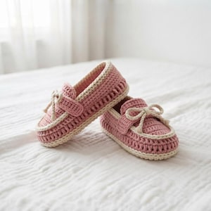 May include: A pair of dusty pink crochet loafers featuring a cream-colored trim along the sole and a decorative bow detail on the top. The shoes are constructed with a textured yarn stitch pattern and have a rounded toe design with a low profile.