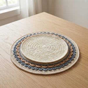 May include: Two round, crocheted placemats stacked on top of each other. The top placemat is solid cream, while the larger bottom placemat features a cream center with concentric rings of scalloped edges in light tan and dusty blue yarn colors.