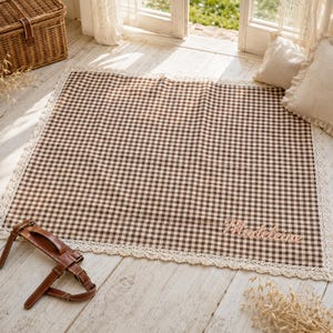 Puede incluir: Una alfombrilla de tela rectangular con un patrón de cuadros vichy en marrón y blanco. La alfombrilla está bordeada por un encaje de ganchillo de color crema. El nombre Madeleine está bordado en cursiva en la esquina inferior derecha.