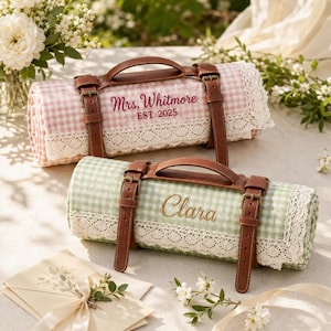 May include: Two rolled picnic blankets with brown leather carrying straps and buckles. One blanket is pink gingham with white lace trim and embroidered text Mrs. Whitmore EST. 2025. The other is green gingham with white lace trim and the name Clara.