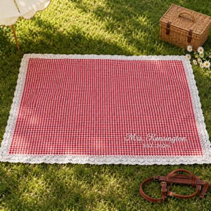May include: A rectangular picnic blanket featuring a red and white gingham pattern with a white lace border. The blanket is personalised with white embroidered text that reads Mrs. Kensington and EST. 2026 in the lower right corner.