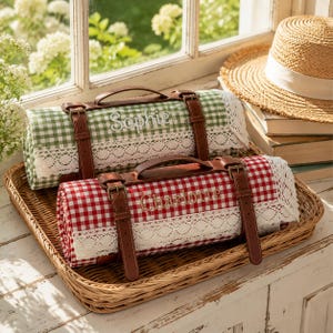 May include: Two rolled picnic blankets with brown leather straps and handles. One blanket features a green and white gingham pattern with the name Sophie embroidered, while the other is red and white gingham with the name Charlotte embroidered.