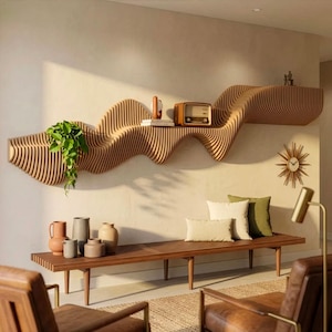 Op de afbeelding: Een lange, golvende wandplank van lichtbruin hout met een lattenontwerp. De plank bevat een groene hangplant, een klein rond object en een radio in vintage-stijl. Daaronder staat een houten bank met diverse keramische vazen en twee kussens.