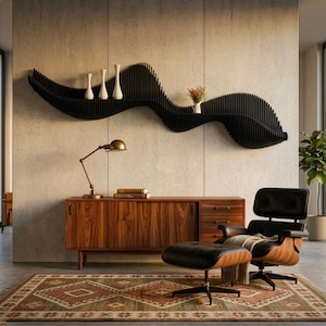 Op de afbeelding: Een zwarte, golfvormige wandplank bevat drie witte vazen en een kleine bloemenvaas. Daaronder staat een houten dressoir met lades en deuren, waarop een messing bureaulamp en boeken staan. Een zwarte leren fauteuil met voetenbank op een patroonkleed.