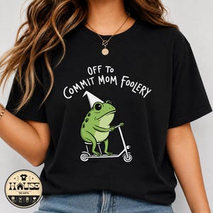 May include: A black short-sleeve t-shirt featuring a graphic of a green frog wearing a white party hat and riding a white scooter. Above the frog, the text reads OFF TO COMMIT MOM FOOLERY in white lettering. The shirt has a crew neckline.