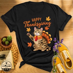 May include: A black short-sleeved t-shirt featuring a graphic of a tabby cat wearing a turkey-feather fan and a red tie. The text "HAPPY Thanksgiving" is printed in orange above the cat, surrounded by orange and yellow autumn leaves.