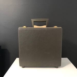May include: A vintage olive green briefcase with a handle and gold-colored clasps. The briefcase has a textured surface and is sitting on a white surface. The handle is gray with a wooden accent.