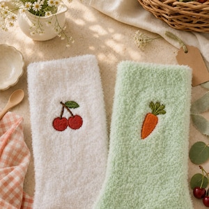 Puede incluir: Un par de calcetines mullidos, uno blanco con un motivo de cerezas rojas bordadas y otro verde claro con un motivo de zanahoria naranja bordada. Los calcetines presentan una textura suave y afelpada sobre una superficie neutra.