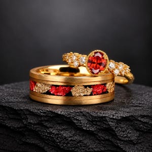 Puede incluir: Un conjunto de anillos en tono dorado que consta de una banda ancha con una incrustación central de fragmentos texturizados rojos y dorados, y un anillo más fino con una piedra roja de corte ovalado rodeada de pequeñas piedras claras.