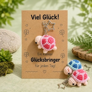 Könnte beinhalten: Ein gehäkelter Schildkröten-Schlüsselanhänger mit einem rosa-weißen Panzer an einem sternförmigen Metallring auf einer beigen Karte. Die Karte trägt den Text "Viel Glück!" und "Ein kleiner Glücksbringer für jeden Tag!" sowie Zeichnungen von vierblättrigen Kleeblättern.
