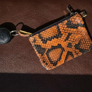 Può includere: Un portamonete rettangolare con un vivace motivo a pelle di serpente arancione e nero e una chiusura a zip nera. Un portachiavi in metallo argentato è fissato al lato del portamonete tramite un piccolo passante in pelle gialla.