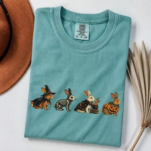 May include: A teal crewneck t-shirt featuring a graphic of four rabbits. One wears a witch hat and cape, one is a skeleton, one wears a star-patterned cape next to a cauldron, and one is inside a pumpkin. The inner label reads Comfort Colors.