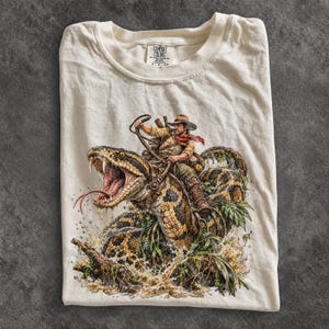 Puede incluir: Una camiseta de manga corta color crema con una ilustración gráfica de una persona con sombrero montando una serpiente grande con la boca abierta sobre el agua. La camiseta tiene una etiqueta de la marca Comfort Colors en el cuello.