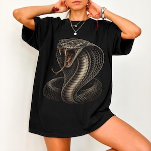 Op de afbeelding: Een zwart t-shirt met korte mouwen en een grote, gedetailleerde grafische print van een cobra met een open bek, met slagtanden en een gespleten tong. De slang is afgebeeld in tinten donkergrijs en bruin met zichtbare schubben en textuur.