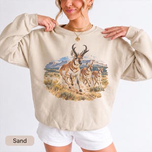 May include: A sand-colored crewneck sweatshirt featuring a graphic print of three pronghorn antelopes running through a desert landscape with mesas in the background. The illustration uses earthy tones of brown, tan, and blue to depict the animals.
