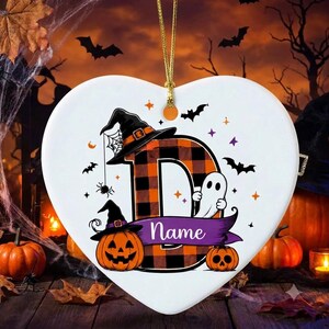 May include: A white heart-shaped ornament featuring a large letter D with an orange and black buffalo plaid pattern. The design includes a witch hat, a ghost, a jack-o'-lantern, and a purple banner with the text Name, suspended by a gold cord.