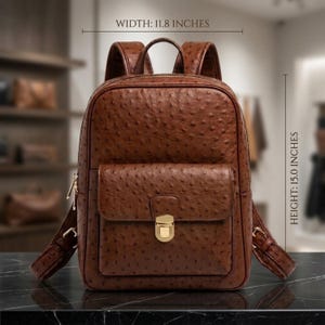 Peut inclure: Un sac à dos marron avec une texture gaufrée façon autruche, doté d'une poche avant avec un fermoir en métal doré, de bretelles réglables et d'une poignée supérieure. Le sac mesure 30 cm de largeur et 38,1 cm de hauteur.