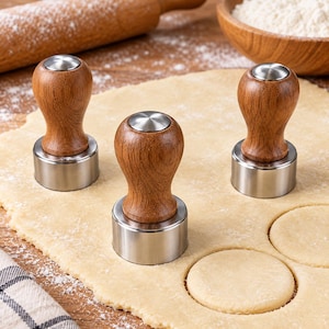 Könnte beinhalten: Drei kreisförmige Teigausstecher mit glatten, hellbraunen Holzgriffen und polierten Edelstahlbasen. Jedes Werkzeug verfügt über einen abgerundeten, ergonomischen Griff und einen scharfen Metallrand zum Ausstechen gleichmäßiger Kreise aus Teig.
