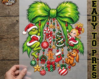 May include: A Christmas-themed DTF transfer sheet featuring a large green bow with hanging ornaments including a green creature in a Santa hat, a blonde girl, a brown dog, a candy cane, a gingerbread man, a stocking, and a gift box.