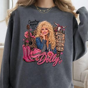 May include: A charcoal gray sweatshirt featuring a graphic print of a blonde woman in a denim jacket, a guitar, pink cowboy boots, and a pink hat. Text includes Country Roads Take Me Home, Jolene, 9 to 5, I Will Always Love You, and Dolly.