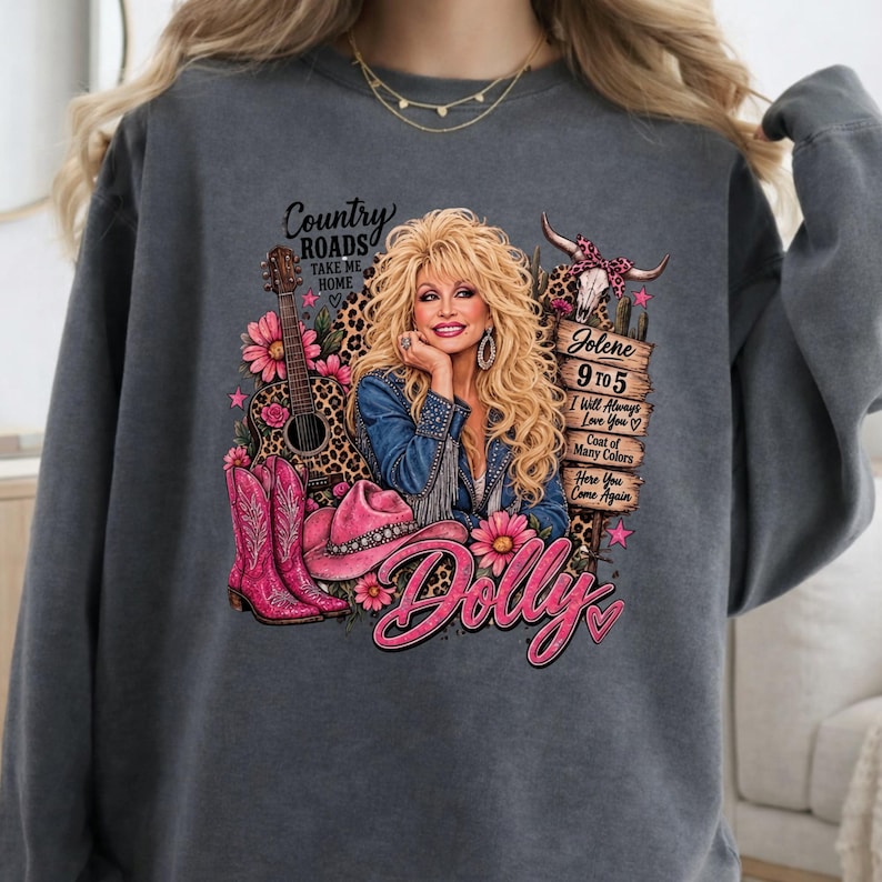 May include: A charcoal gray sweatshirt featuring a graphic print of a blonde woman in a denim jacket, a guitar, pink cowboy boots, and a pink hat. Text includes Country Roads Take Me Home, Jolene, 9 to 5, I Will Always Love You, and Dolly.