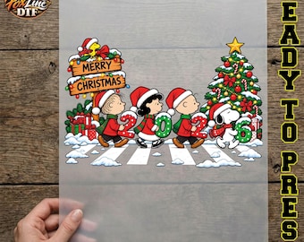 May include: A DTF transfer sheet featuring a Peanuts holiday graphic with Linus, Lucy, Charlie Brown, and Snoopy wearing Santa hats. They carry the numbers 2, 0, 2, 6 in front of a decorated Christmas tree and a Merry Christmas sign.
