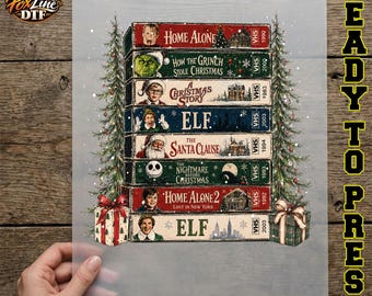 May include: A stack of eight VHS tapes featuring Christmas movie titles including Home Alone, How the Grinch Stole Christmas, A Christmas Story, Elf, The Santa Clause, The Nightmare Before Christmas, Home Alone 2, and Elf, flanked by two pine trees.