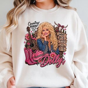 May include: A cream-colored sweatshirt featuring a graphic print of Dolly Parton with blonde hair, a denim jacket, and a guitar. The design includes pink cowboy boots, a pink hat, flowers, a cow skull, and text reading Country Roads, Jolene, 9 to 5.