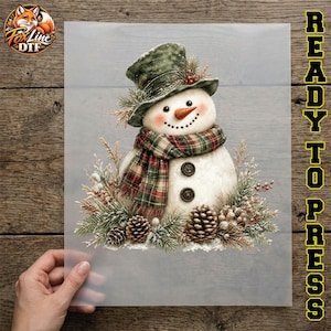 May include: A snowman graphic transfer sheet featuring a snowman wearing a green top hat and a red and green plaid scarf. The snowman has a carrot nose, two dark buttons, and is surrounded by pine branches, pinecones, and red berries.