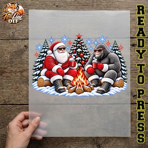 May include: A DTF transfer sheet featuring a graphic of Santa Claus and a Bigfoot sitting by a campfire in the snow. Both figures wear sunglasses and hold red mugs. A decorated Christmas tree stands behind them with blue snowflakes.