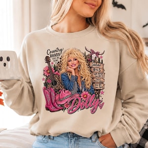 May include: A beige crewneck sweatshirt featuring a graphic print of a blonde woman, a guitar, pink cowboy boots, and a pink hat. The text includes Country Roads Take Me Home, Jolene, 9 to 5, I Will Always Love You, Coat of Many Colors, Here You Come Again, and Dolly.
