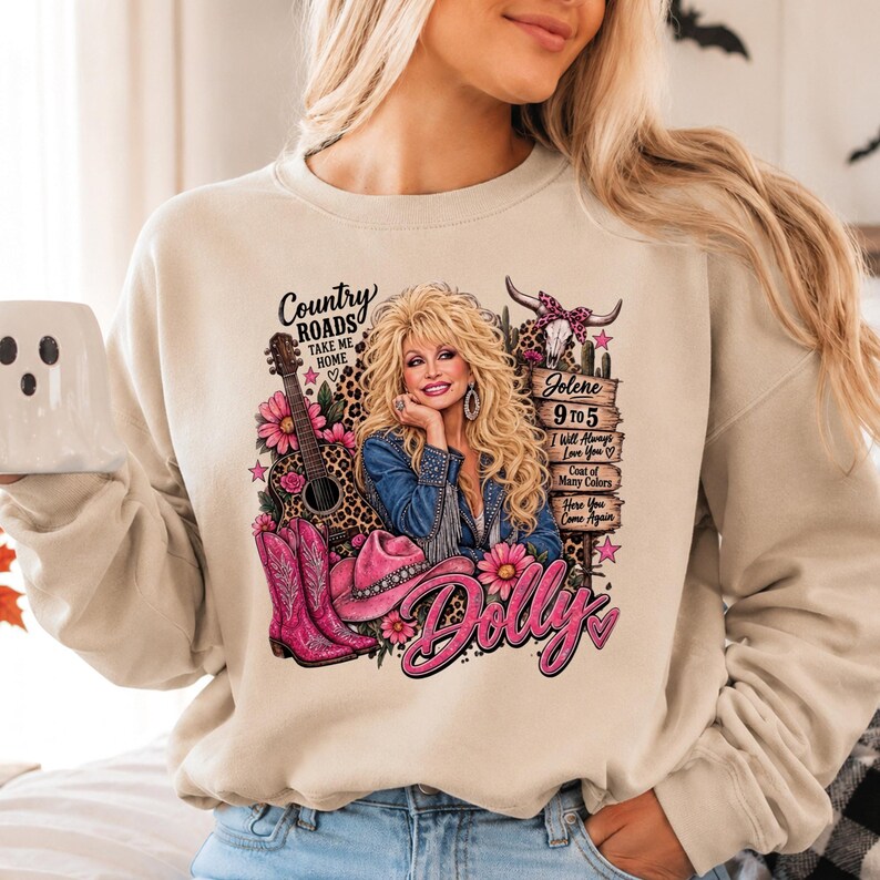 May include: A beige crewneck sweatshirt featuring a graphic print of a blonde woman, a guitar, pink cowboy boots, and a pink hat. The text includes Country Roads Take Me Home, Jolene, 9 to 5, I Will Always Love You, Coat of Many Colors, Here You Come Again, and Dolly.